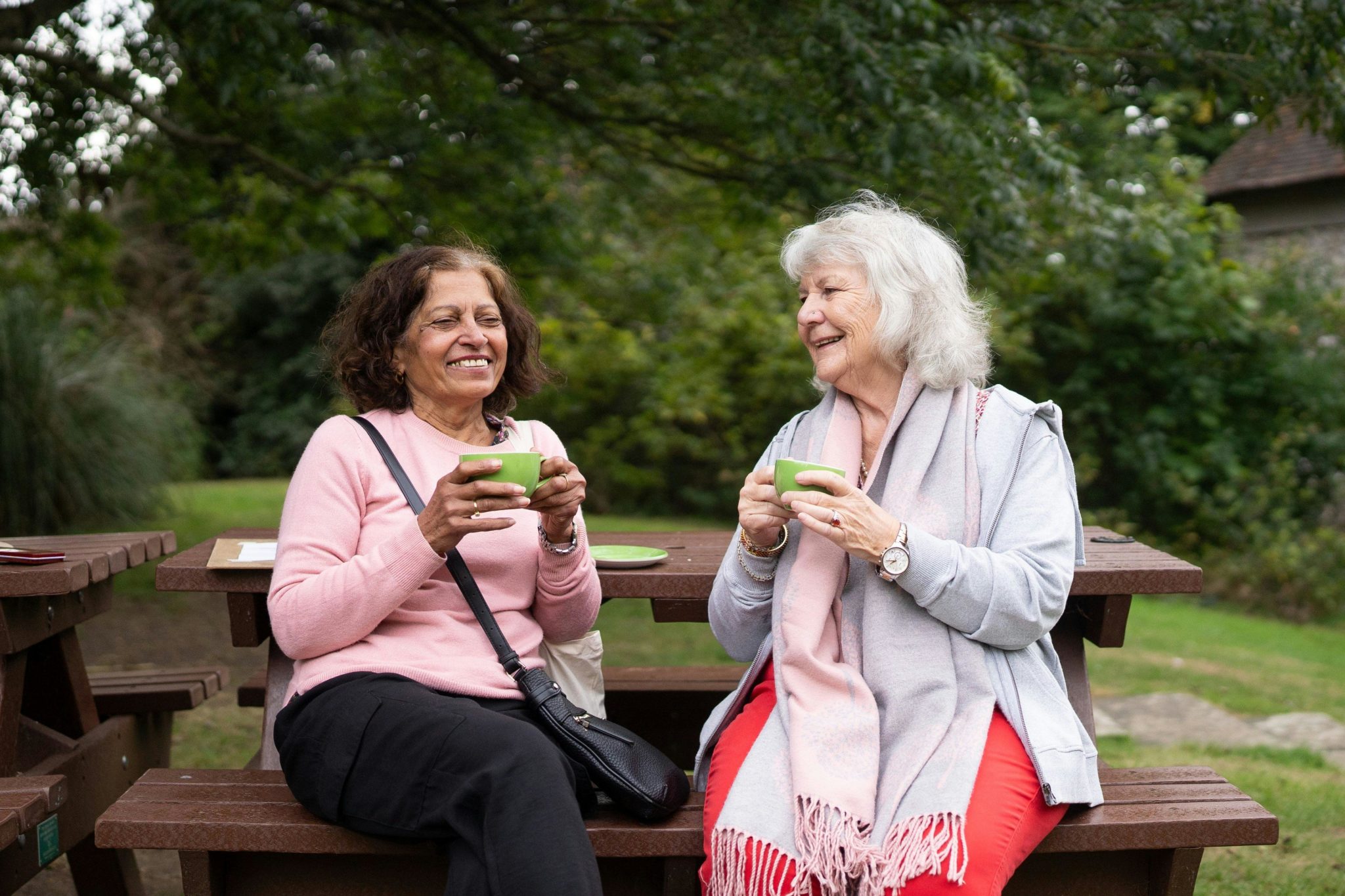 Two women smiling and holding cups on a park bench