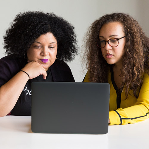 Two people sit at a table in front of a laptop, one with hands on the keyboard, instructing, while the other watches