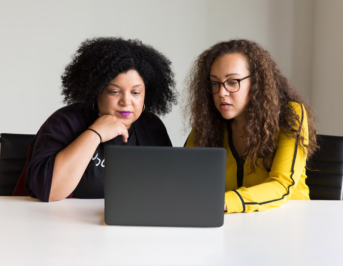 Two people sit at a table in front of a laptop, one with hands on the keyboard, instructing, while the other watches