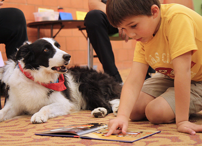 A child sits on the ground, finger pointing at an open book, while a dog sits next to him watching