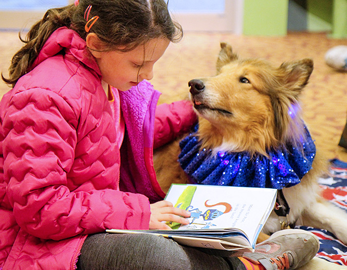 a child sits on the ground reading a book and petting a dog which looks up at her