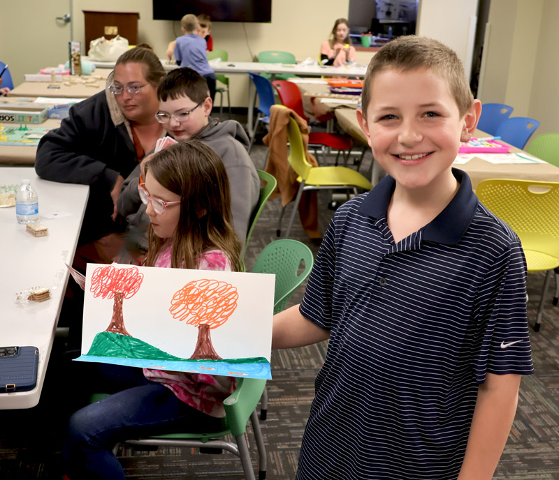 a child holds up a drawing he made while other children and parents do crafts at tables in the background