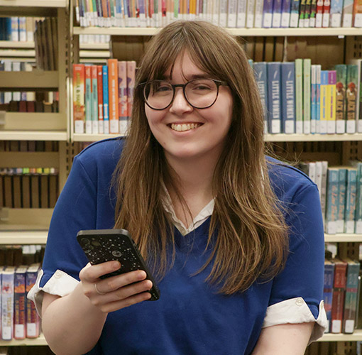 a person sitting in the library in front of a shelf of books holds up their phone in a reading position while smiling at the camera